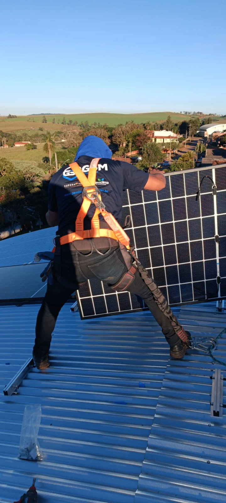 Profissional com capacete e cinturão manuseando módulo fotovoltaico sobre cobertura metálica