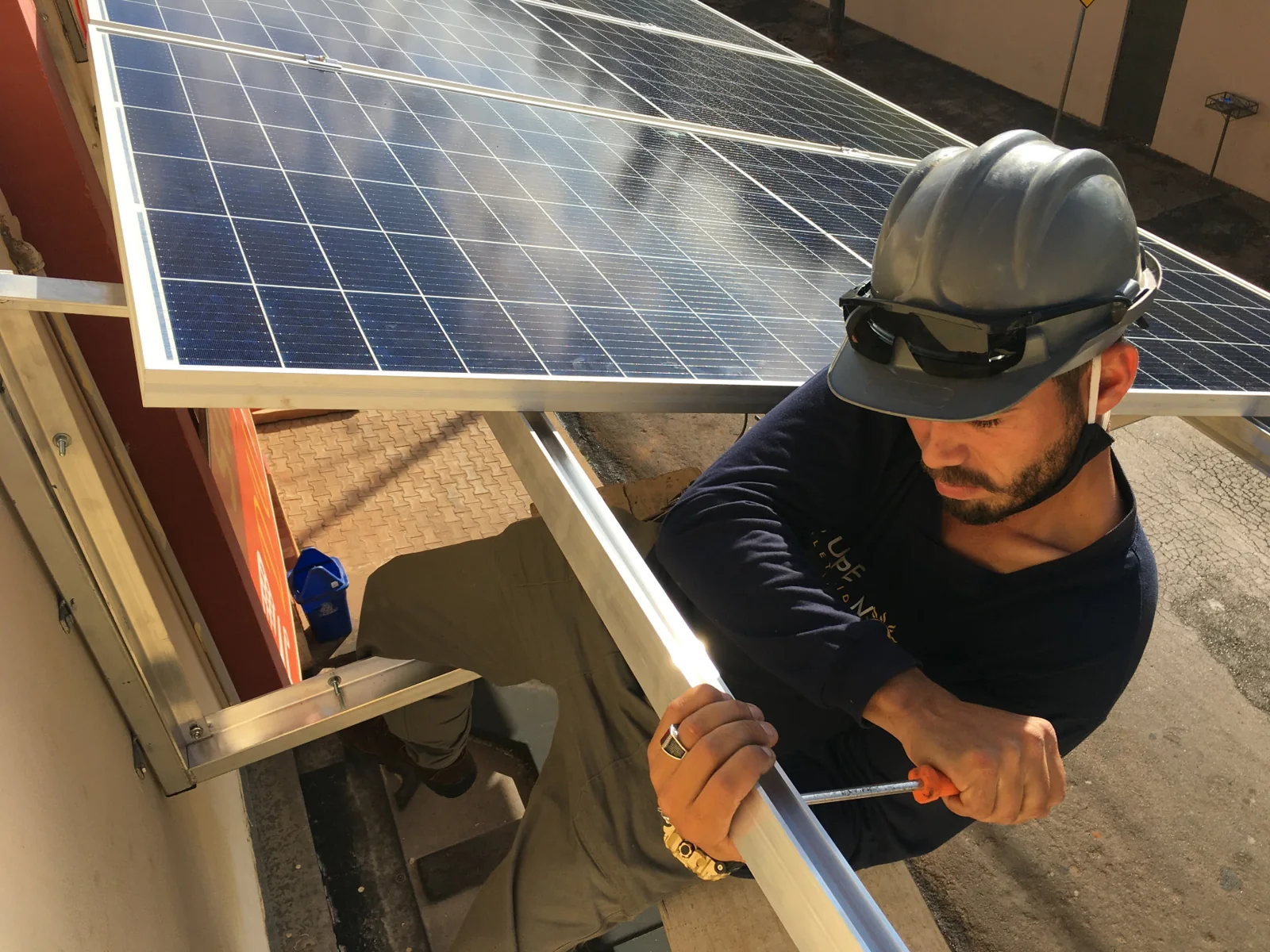 Profissional com capacete e óculos de proteção segurando a estrutura de um módulo fotovoltaico junto à cobertura